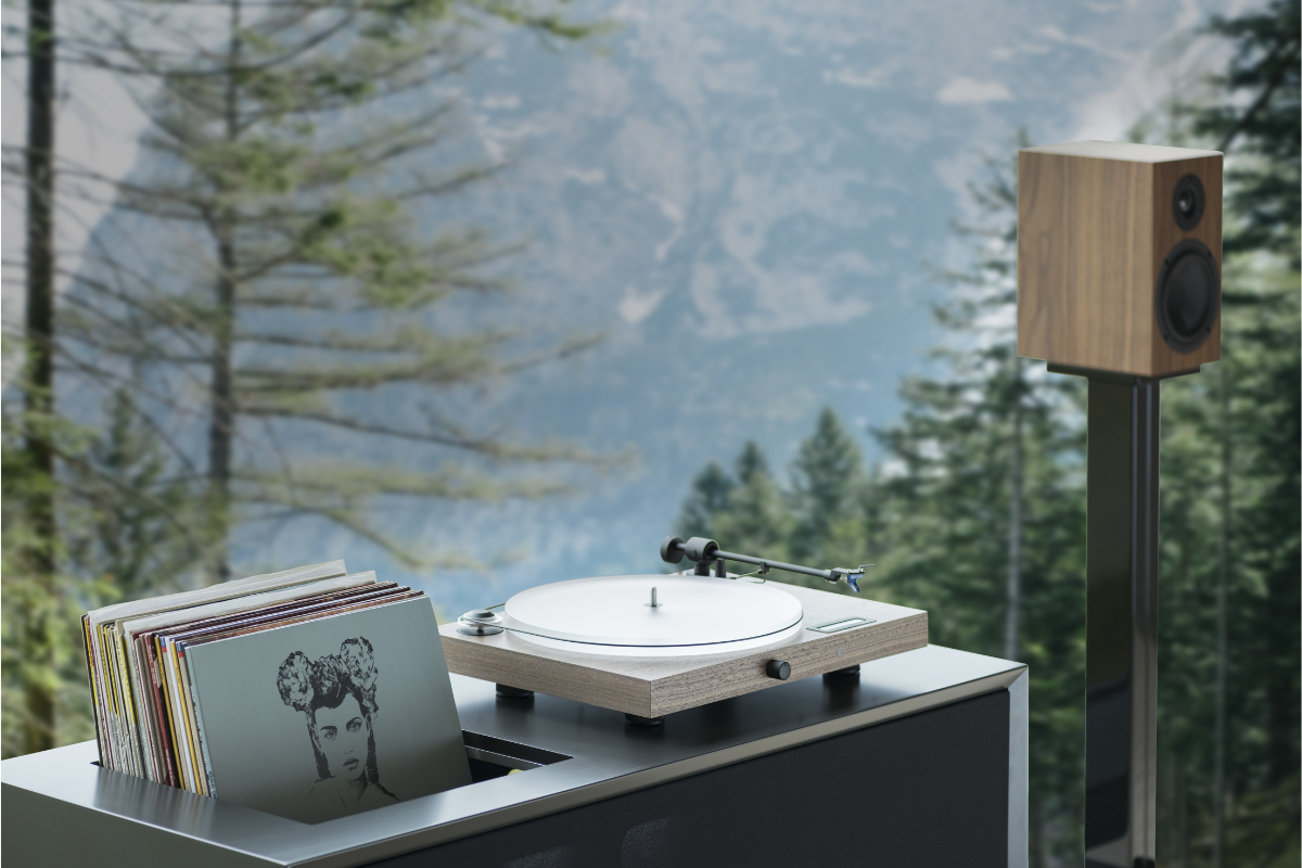 Pro-Ject Plattenspieler in Holzoptik mit Lautsprecher und Schallplatten, stimmungsvolle HiFi-Szene vor alpiner Bergkulisse