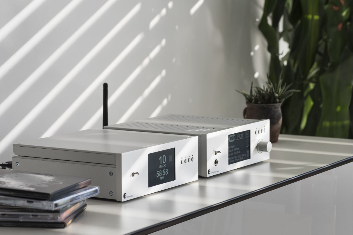 Zwei silberne HiFi-Komponenten mit Displays auf einem Sideboard im Sonnenlicht, daneben ein Stapel CDs und eine Zimmerpflanze.