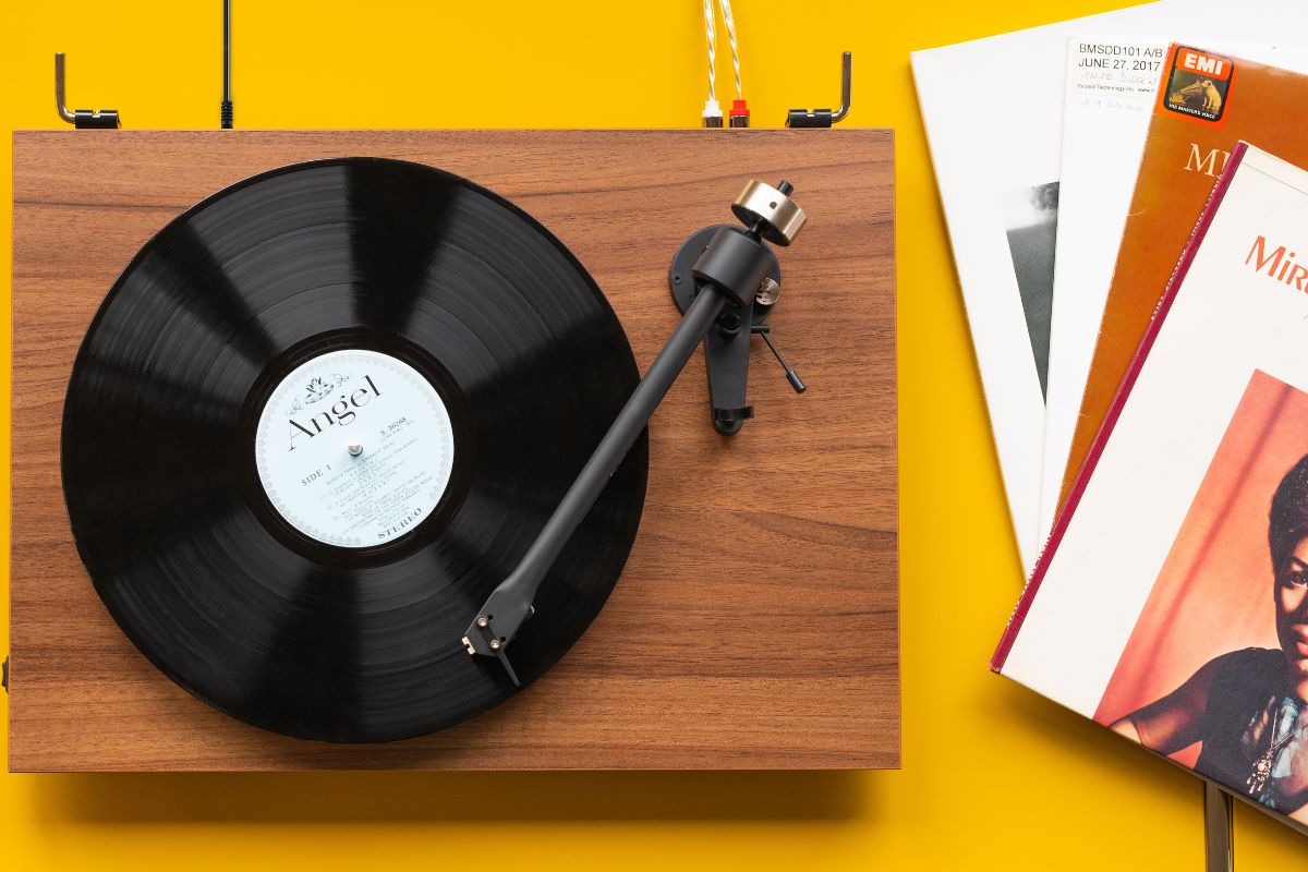 Plattenspieler mit Holzgehäuse und schwarzer Schallplatte auf gelbem Hintergrund, daneben liegen mehrere Schallplattenhüllen.