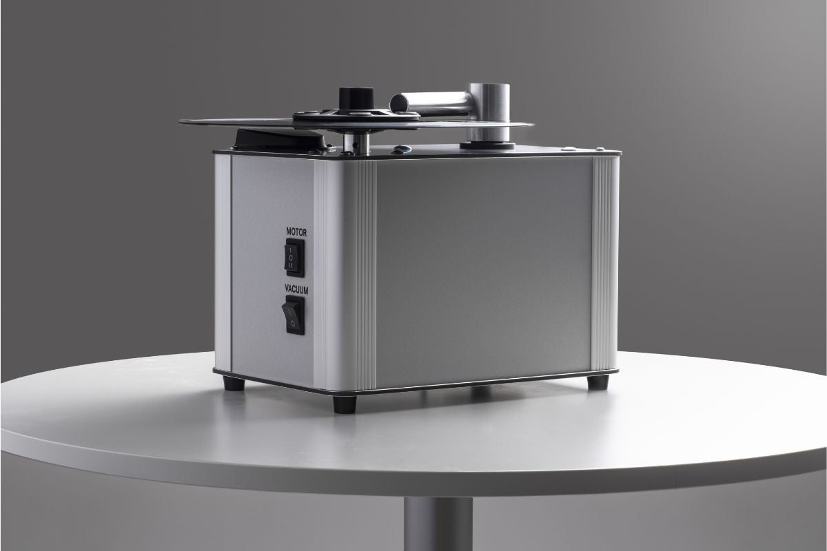 Plattenwaschmaschine in silbernem Gehäuse auf rundem Tisch, mit Schaltern für Motor und Vakuum sowie Absaugarm oben.