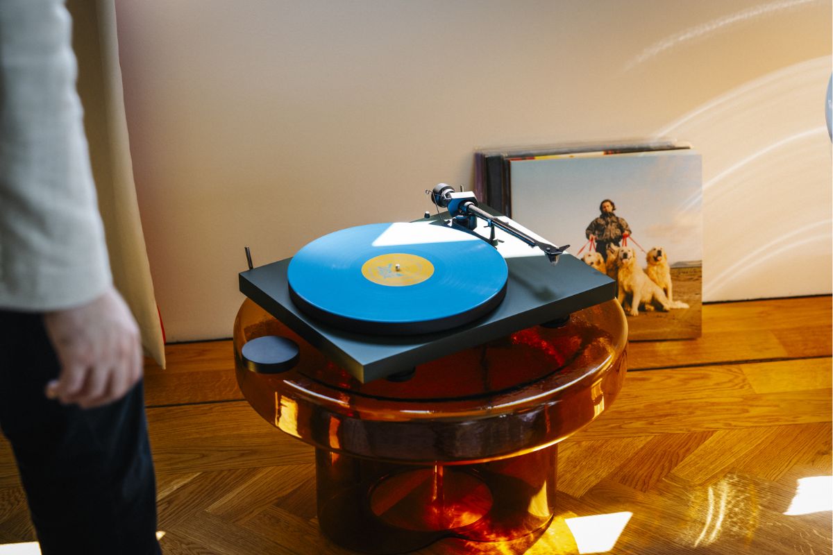 Plattenspieler mit blauer Schallplatte auf einem runden, transparenten Tisch, im Hintergrund ein Plattencover mit einer Person und zwei Hunden.