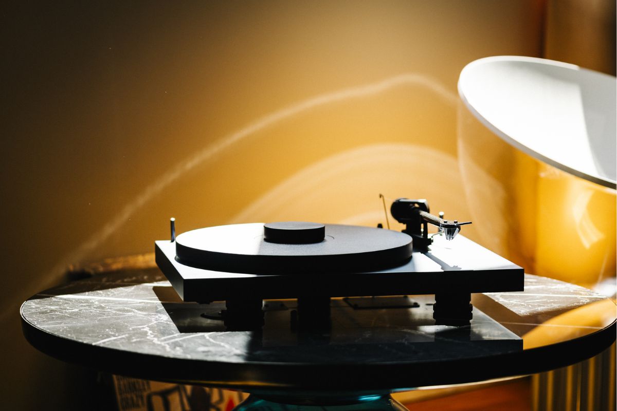 Ein schwarzer Plattenspieler steht auf einem runden Marmortisch in warmem Sonnenlicht. Der Tonarm befindet sich rechts, der Plattenteller ist leer, und im Hintergrund fällt goldenes Licht auf eine gelbe Wand und eine moderne Lampe.