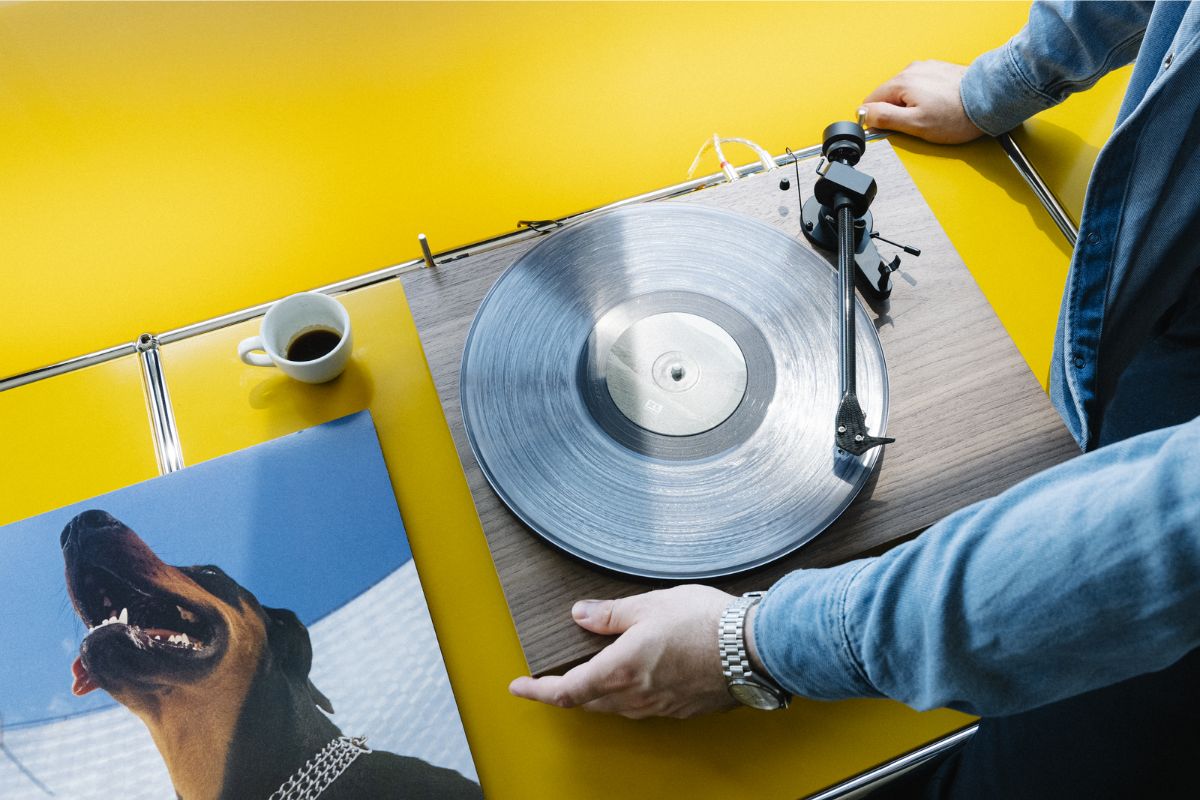 Person bedient einen Plattenspieler mit transparenter Vinyl-Schallplatte auf gelbem Sideboard, daneben eine Espressotasse und ein Albumcover mit einem Hund.
