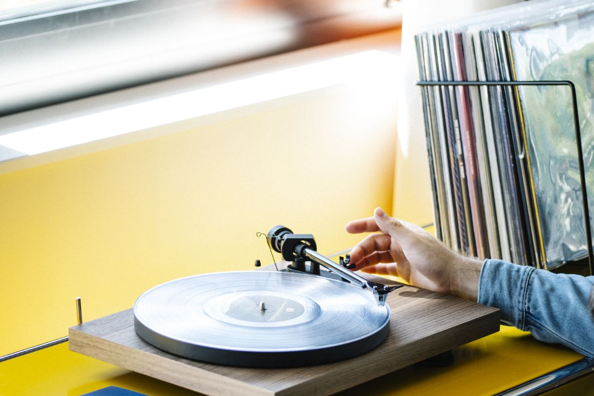 Hand legt eine transparente Vinyl-Schallplatte auf einen Holzplattenspieler, im Hintergrund steht eine geordnete Sammlung von Schallplatten