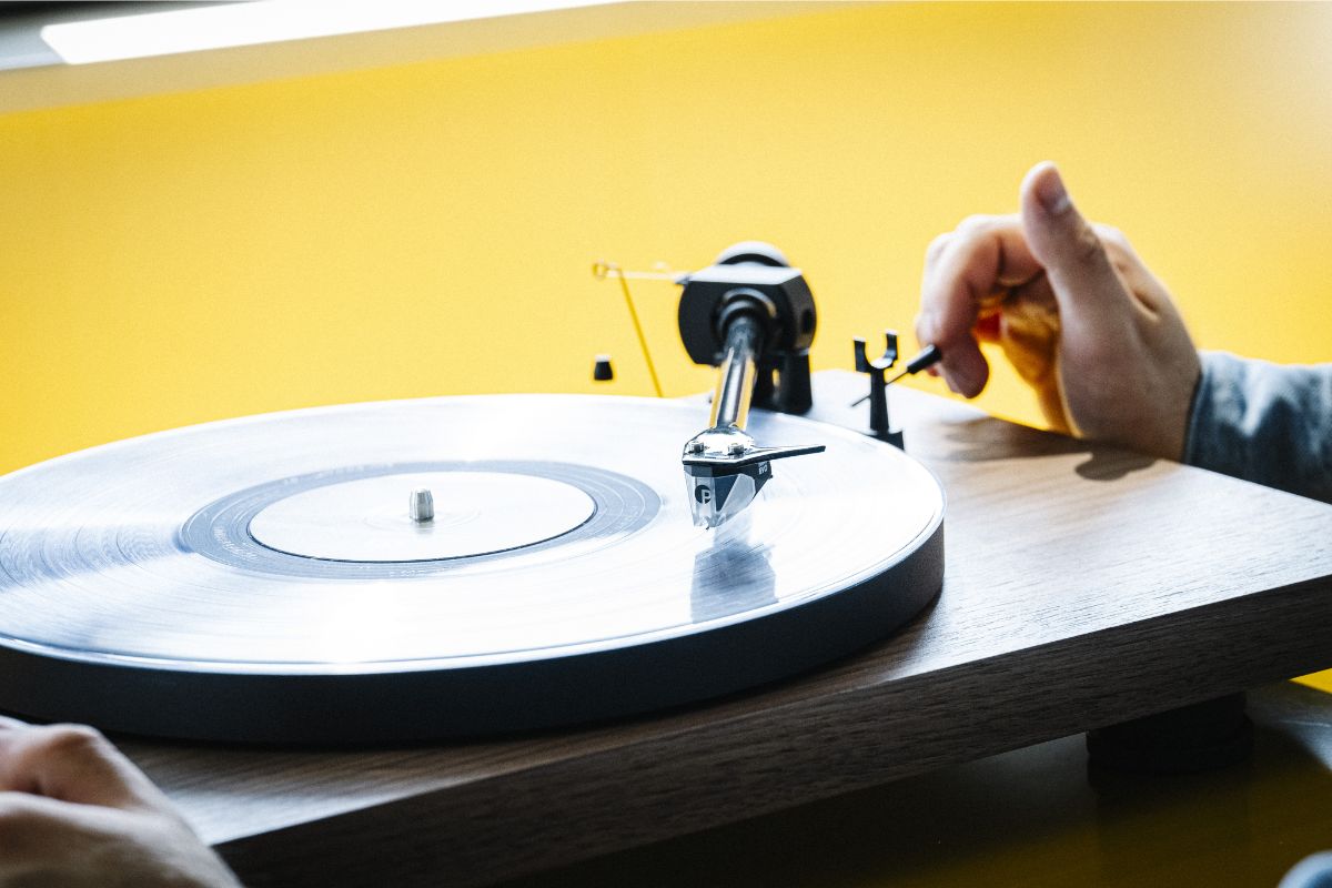 Hand justiert den Tonarm eines Pro-Ject Plattenspielers mit transparenter Nadel, vor gelbem Hintergrund
